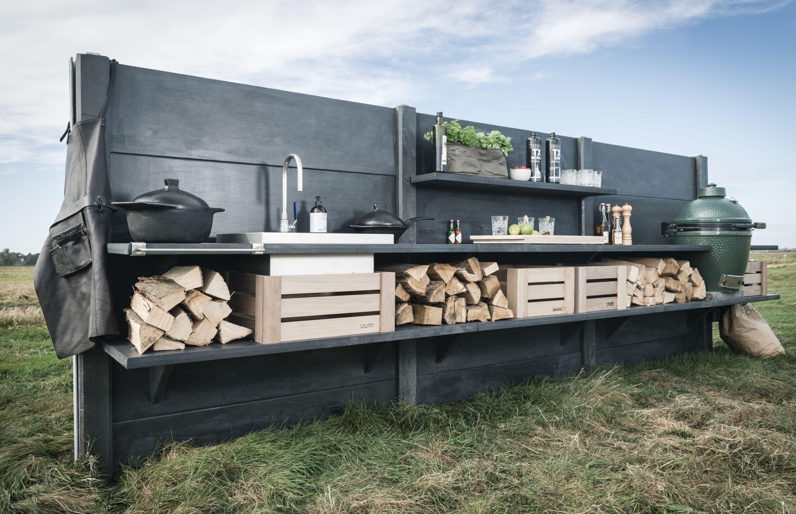 Antracite concrete outdoor kitchen stocked with wood and a green egg standing in a grass field